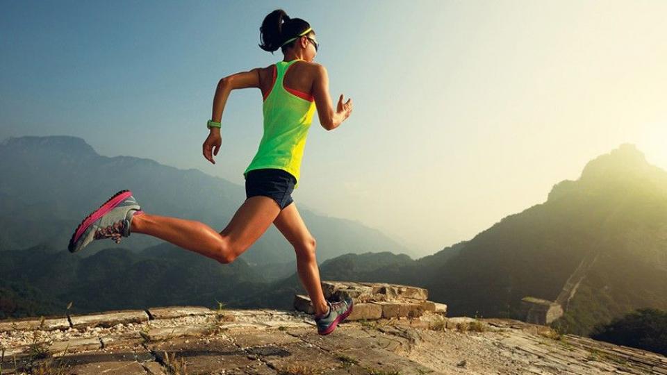 A person in athletic gear runs along a rugged, mountainous path with a hazy, expansive landscape in the background.